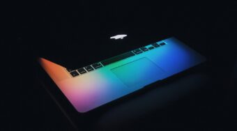 An apple laptop with its screen half closed and multi-coloured light shining on the keyboard.